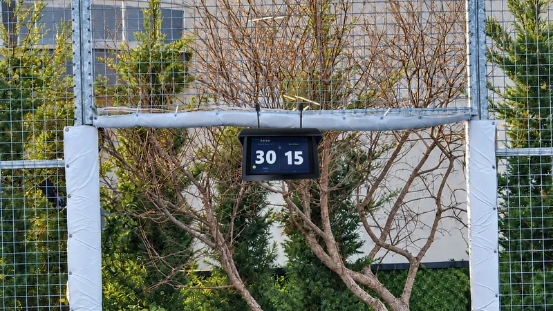 Padel kortunun giriş kapısının üst kirişinde, yağmur korumalı tutucunun içinde kort tableti: skor 30-15